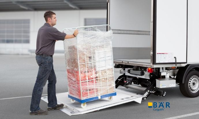 Un uomo spinge un contenitore su ruote sulla piattaforma di una sponda montacarichi BAR montata su un furgone