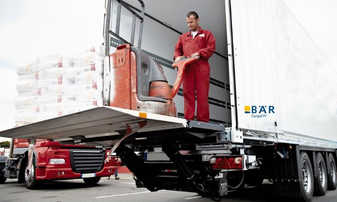 Autista con transpallet elettrico e pallet pesante sulla piattaforma di una piattaforma elevatrice Bar retrattile