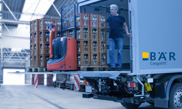 Il conducente si trova con diverse pallet di bevande e un transpallet elettrico sulla piattaforma di una sponda montacarichi BAR