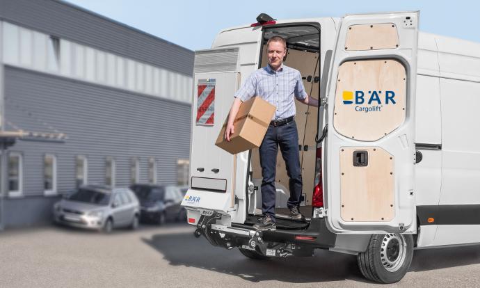 Un uomo scende dal furgone con un pacco attraverso la porta posteriore, mentre il Bar VanLift FreeAccess rimane fermo.