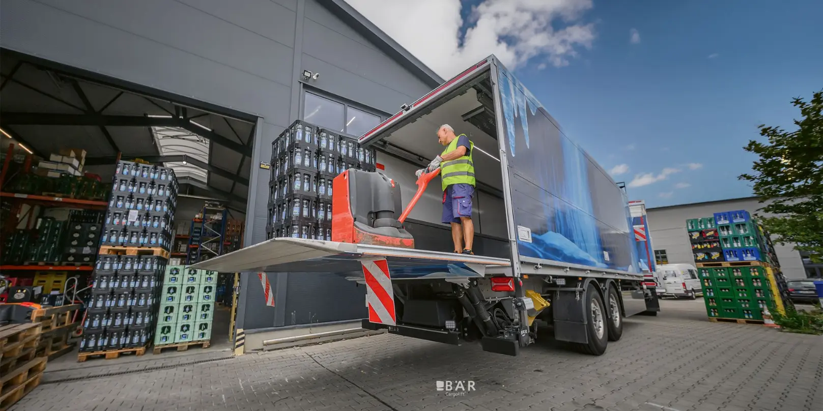 Un uomo scarica un pallet di bevande con transpallet tramite una sponda caricatrice Bar Cargolift montata sul rimorchio di un autotreno